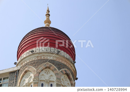 A low-angle shot featuring the iconic red dome of the Taj Mahal Palace Hotel in Mumbai showcasing its intricate Indo-Saracenic architectural details against a clear pale blue sky. A low-angle shot featuring the iconic red dome of the Taj Mahal Palace Hotel in Mumbai showcasing its intricate Indo-Saracenic architectural details against a clear pale blue sky. 140728994