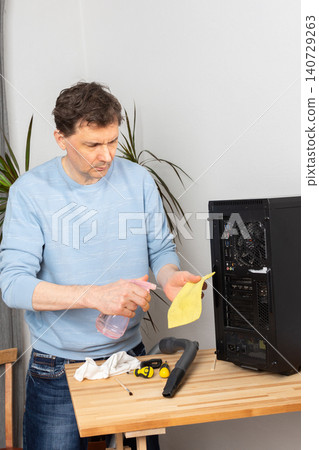 A 53-year-old man removes dust from a computer system unit while standing at a wooden table. He holds a yellow sticky note and spray cleaner in his hands A 53-year-old man removes dust from a computer system unit while standing at a wooden table. He holds a yellow sticky note and spray cleaner in his hands 140729263