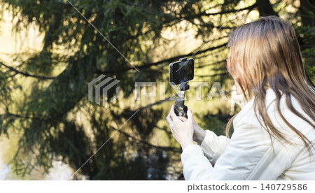 Woman filming with smartphone on a gimbal in a forest. Outdoor content creation gear for travel vlogging. Sunlit woodland scene for social media video production.. Woman filming with smartphone on a gimbal in a forest. Outdoor content creation gear for travel vlogging. Sunlit woodland scene for social media video production.. 140729586