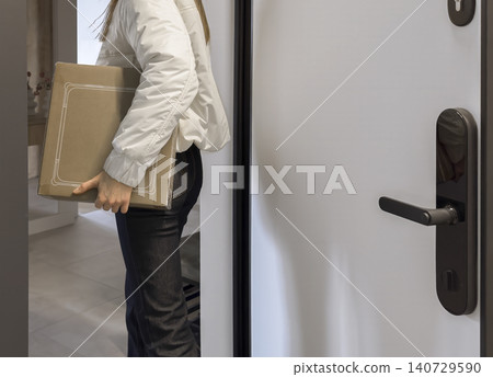 Woman carrying a cardboard box enters a home doorway. Indoor delivery and moving in a modern apartment. Household logistics and unpacking at the entrance.. Woman carrying a cardboard box enters a home doorway. Indoor delivery and moving in a modern apartment. Household logistics and unpacking at the entrance.. 140729590
