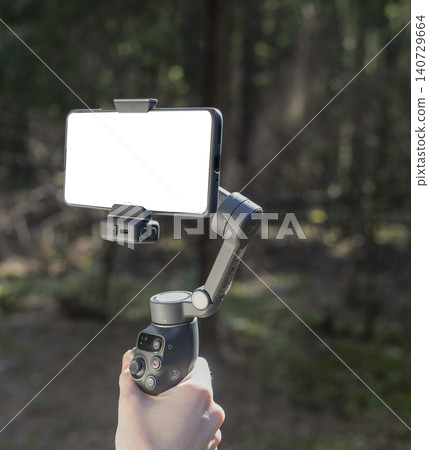 Smartphone on a gimbal stabilizer in a forest. Mobile filming setup with handheld support and blurred woodland background. Outdoor content creation for video recording and social 140729664