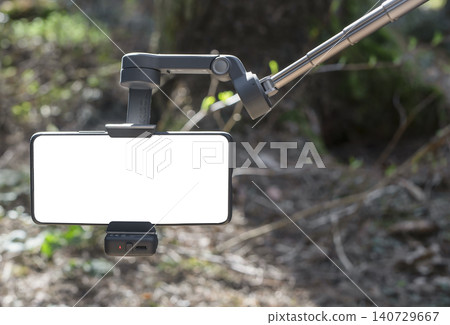 Smartphone on a gimbal stabilizer in a forest. Blank screen mobile filming gear hangs outdoors with blurred woodland background. Content creation setup for video recording and Smartphone on a gimbal stabilizer in a forest. Blank screen mobile filming gear hangs outdoors with blurred woodland background. Content creation setup for video recording and 140729667