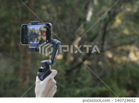 Smartphone mounted on a handheld gimbal in a forest. Mobile video recording setup for outdoor content creation. Travel vlogging gear with nature background.. Smartphone mounted on a handheld gimbal in a forest. Mobile video recording setup for outdoor content creation. Travel vlogging gear with nature background.. 140729682