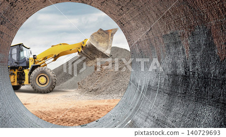 A loader lifts gravel at a construction site. The scene is viewed through a large pipe. The sky is cloudy and the ground is dusty with construction materials A loader lifts gravel at a construction site. The scene is viewed through a large pipe. The sky is cloudy and the ground is dusty with construction materials 140729693
