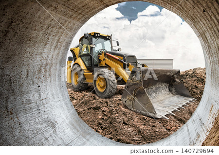 A powerful excavator-loader moves earth and debris at a construction site, as seen from a large chimney. The sky above the machinery and equipment is partly cloudy A powerful excavator-loader moves earth and debris at a construction site, as seen from a large chimney. The sky above the machinery and equipment is partly cloudy 140729694