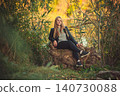 Young blonde woman in leather jacket sitting on tree stump in autumn forest 140730088