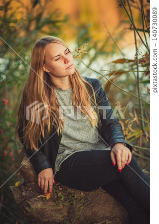 Contemplative blonde woman in leather jacket relaxing outdoors in autumn park Contemplative blonde woman in leather jacket relaxing outdoors in autumn park 140730089