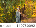 Young blonde woman in grey coat walking park path enjoying golden autumn light 140730092