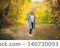 Blonde woman walking on autumn park path. Casual female in grey coat enjoys golden nature scenery 140730093