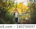 Blonde woman in grey coat stands by water, surrounded by lush autumn foliage, thoughtful expression, natural light 140730097