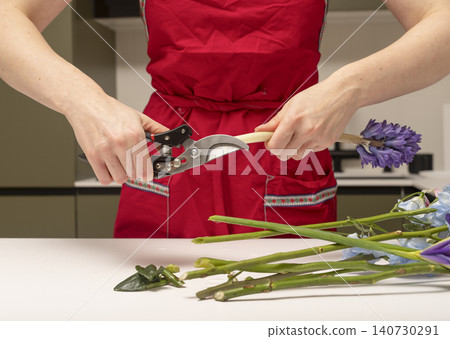 Florist cutting flower stem with pruning shears at table. Hands trimming plant preparing floral material for arrangement. Concept of professional flower preparation process.. Florist cutting flower stem with pruning shears at table. Hands trimming plant preparing floral material for arrangement. Concept of professional flower preparation process.. 140730291