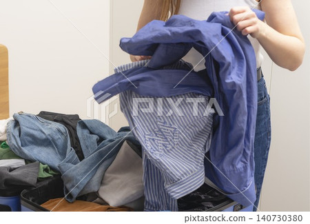 Person holding pile of clothes in messy packing process for suitcase. Hands prepare garments without order before placing into luggage. Concept of travel preparation chaos organization.. Person holding pile of clothes in messy packing process for suitcase. Hands prepare garments without order before placing into luggage. Concept of travel preparation chaos organization.. 140730380