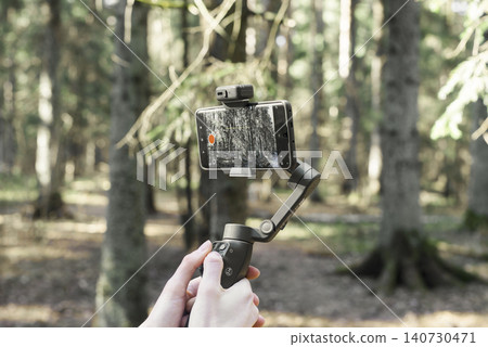 Smartphone mounted on a gimbal in a forest. Handheld stabilizer supports mobile filming in nature. Outdoor content creation gear for travel and vlogging.. 140730471