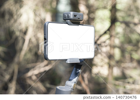 Smartphone mounted on a gimbal in a forest setting. Mobile filming gear stands against a blurred woodland background. Outdoor content creation and videography concept.. Smartphone mounted on a gimbal in a forest setting. Mobile filming gear stands against a blurred woodland background. Outdoor content creation and videography concept.. 140730476