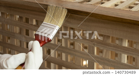 Paintbrush applies finish to an acacia wood lattice table. Gloved hand coats the slatted surface during furniture maintenance. DIY woodwork and home improvement concept.. Paintbrush applies finish to an acacia wood lattice table. Gloved hand coats the slatted surface during furniture maintenance. DIY woodwork and home improvement concept.. 140730489