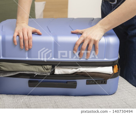 Woman pressing suitcase lid to close overfilled luggage during packing. Hands apply force to compress contents inside case. Concept of travel preparation overload.. Woman pressing suitcase lid to close overfilled luggage during packing. Hands apply force to compress contents inside case. Concept of travel preparation overload.. 140730494