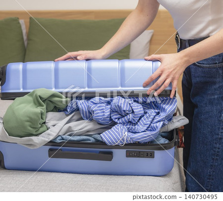 Woman struggling to close overfilled suitcase at home. Clothes exceed luggage capacity during travel preparation. Concept of packing overload and travel stress.. Woman struggling to close overfilled suitcase at home. Clothes exceed luggage capacity during travel preparation. Concept of packing overload and travel stress.. 140730495