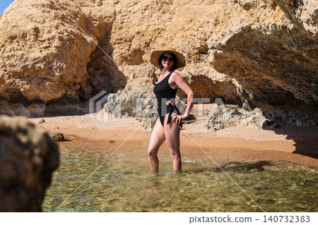 Young woman in a black swimsuit, hat, and sunglasses posing for the photographer against a stunning rocky seaside backdrop, capturing the essence of summer and beach vibes Young woman in a black swimsuit, hat, and sunglasses posing for the photographer against a stunning rocky seaside backdrop, capturing the essence of summer and beach vibes 140732383