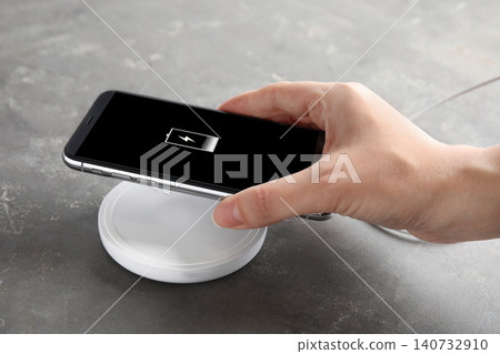 Woman putting mobile phone onto wireless charger at grey table, closeup Woman putting mobile phone onto wireless charger at grey table, closeup 140732910