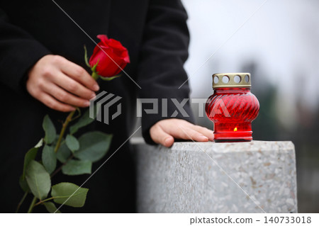 Woman holding red rose near grey granite tombstone with candle outdoors, closeup. Funeral ceremony Woman holding red rose near grey granite tombstone with candle outdoors, closeup. Funeral ceremony 140733018