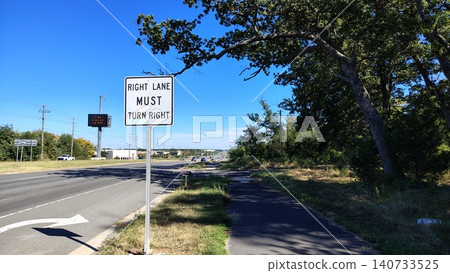 In the foreground, a white road sign reads, "Must turn right in right lane." In the distance, an electronic road sign rises from a tall pole above the highway. In the foreground, a white road sign reads, "Must turn right in right lane." In the distance, an electronic road sign rises from a tall pole above the highway. 140733525