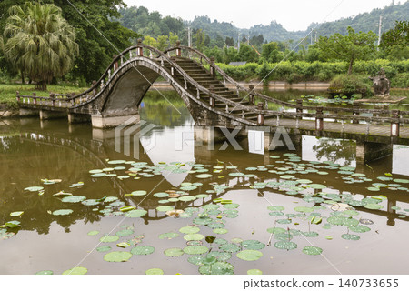 In Dazu Lotus Manor, a steeply stepped stone arch bridge vaults over a calm pond dotted with floating lotus foliage. Its high-arched silhouette casts a clear reflection onto the murky water, while a In Dazu Lotus Manor, a steeply stepped stone arch bridge vaults over a calm pond dotted with floating lotus foliage. Its high-arched silhouette casts a clear reflection onto the murky water, while a 140733655