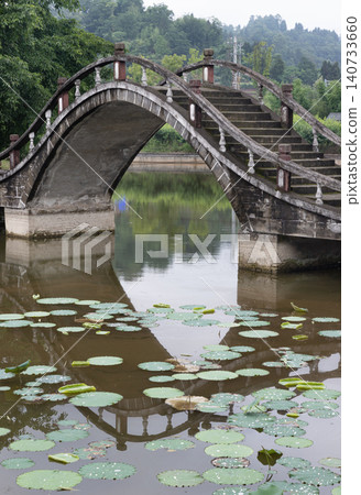 a traditional stone bridge rising into a dramatic, steeply stepped arch over a calm pond, with green lotus leaves drifting on the water's surface below. a traditional stone bridge rising into a dramatic, steeply stepped arch over a calm pond, with green lotus leaves drifting on the water's surface below. 140733660