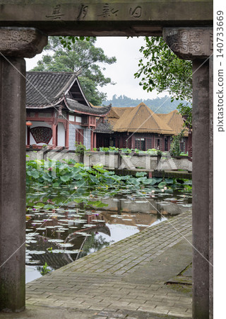 Framed by an inscribed stone archway, a scenic view of Dazu Lotus Manor unfolds. A paved pathway leads to a tranquil pond filled with lush lotus leaves and delicate blossoms. In the background, Framed by an inscribed stone archway, a scenic view of Dazu Lotus Manor unfolds. A paved pathway leads to a tranquil pond filled with lush lotus leaves and delicate blossoms. In the background, 140733669