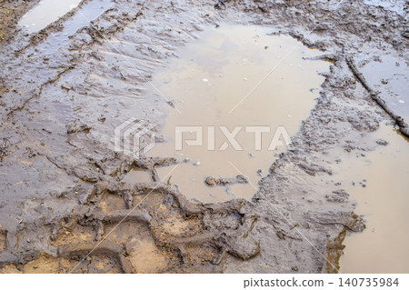 Muddy puddle with tractor tire tracks on winding dirt road, country lane lane with wet clay potholes landscape 140735984