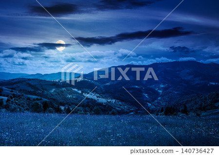 mountain landscape with grassy field at night. full moon over trees on the rolling hills and rural valley beneath a dark sky in the background. mysterious view of carpathian mountains in summer mountain landscape with grassy field at night. full moon over trees on the rolling hills and rural valley beneath a dark sky in the background. mysterious view of carpathian mountains in summer 140736427