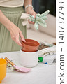 Woman prepares plant pot for repotting, placing container inside a planter with gloves and tools nearby 140737793