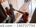 Woman paints wall with roller beside mirror, with dog and ladder visible in room 140737794