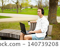 Enjoying remote work and freelancing, a smiling man is working on a laptop outdoors in a sunny park, sitting on a bench with coffee and headphones. 140739208