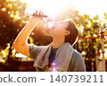A thirsty man is drinking water outdoors after a fitness workout. Covered in sweat with a towel around his neck, he cools down under the bright sun. 140739211