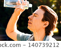 Overcome by summer heat, a man outdoors is pouring water on his face. Cooling down his sweat after a heavy fitness workout, he uses a bottle for refreshment. 140739212