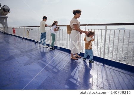 Mother and three children stand at a ferry railing watching the ocean, sharing a quiet family travel moment on deck with light reflections and relaxed summer mood. Mother and three children stand at a ferry railing watching the ocean, sharing a quiet family travel moment on deck with light reflections and relaxed summer mood. 140739215
