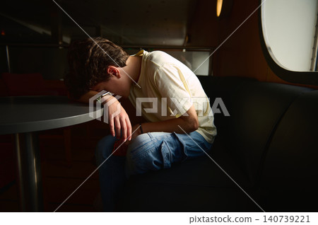 Teenage boy exhausted and emotional, sitting curled on a sofa with head resting on a table near a porthole in a dim ship cabin, conveying fatigue, loneliness and travel weariness. Teenage boy exhausted and emotional, sitting curled on a sofa with head resting on a table near a porthole in a dim ship cabin, conveying fatigue, loneliness and travel weariness. 140739221