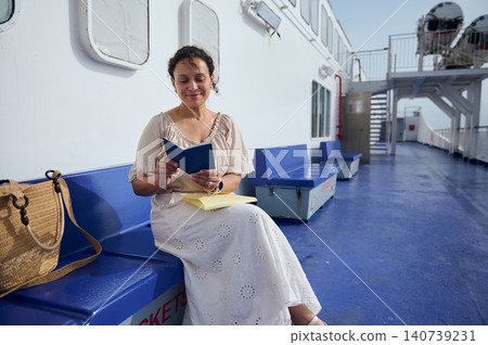 Woman sits on a ferry deck reading a small book with a notebook and tote bag nearby, enjoying a peaceful sunny travel moment on blue benches and open sea in the background. Woman sits on a ferry deck reading a small book with a notebook and tote bag nearby, enjoying a peaceful sunny travel moment on blue benches and open sea in the background. 140739231