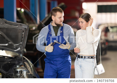 Mechanic advises young woman client on repairing under hood of car in car service station Mechanic advises young woman client on repairing under hood of car in car service station 140739708