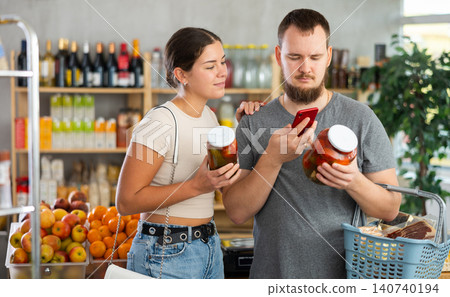 Portrait of man and woman checking the expiration date of canned tomatos and cucumbers using smartphone 140740194