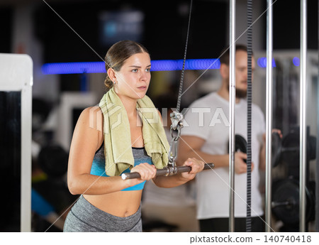 Girl in gym performs exercises for arm muscles using cable crossover machine and pull weighted rope Girl in gym performs exercises for arm muscles using cable crossover machine and pull weighted rope 140740418