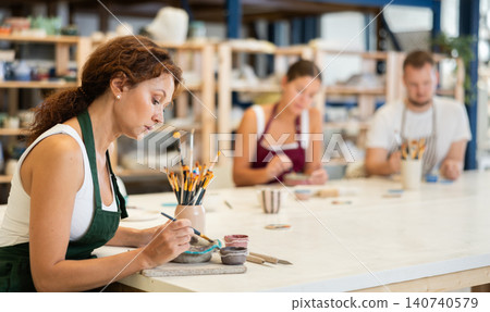 Woman learns to make designs on clay plate using brush and paint in pottery workshop 140740579
