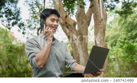 East Asian man talking on phone with laptop for remote work outdoors East Asian man talking on phone with laptop for remote work outdoors 140741490