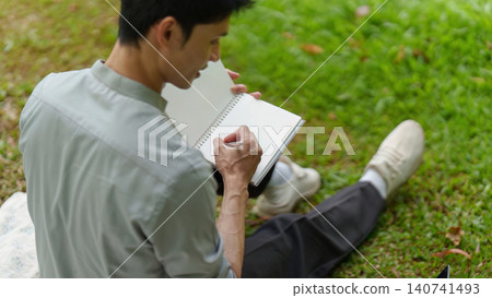 Asian man writing in notebook with pen in park for journaling Asian man writing in notebook with pen in park for journaling 140741493