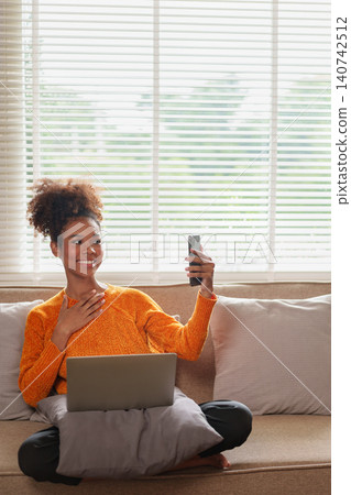 Cheerful African American woman video calling on smartphone while sitting on couch with laptop. Cheerful African American woman video calling on smartphone while sitting on couch with laptop. 140742512