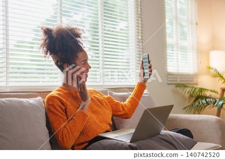 Cheerful African American woman video calling on smartphone while sitting on couch with laptop. Cheerful African American woman video calling on smartphone while sitting on couch with laptop. 140742520