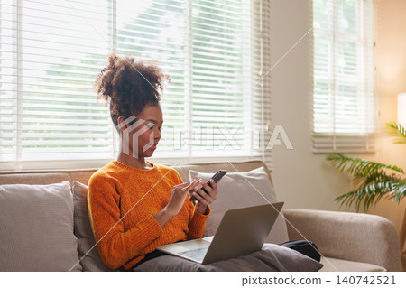 African American woman texting on smartphone while sitting on couch with laptop open, in cozy room. African American woman texting on smartphone while sitting on couch with laptop open, in cozy room. 140742521