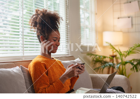 African American woman texting on smartphone while sitting on couch with laptop open, in cozy room. African American woman texting on smartphone while sitting on couch with laptop open, in cozy room. 140742522