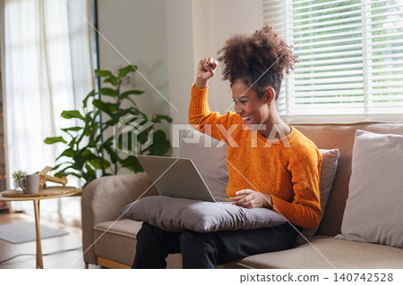 Young black woman celebrating success while working on laptop in cozy living room. 140742528