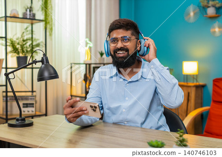 Indian man lifts headphones smiles delighted after audio story on smartphone amazed reaction indoors Indian man lifts headphones smiles delighted after audio story on smartphone amazed reaction indoors 140746103